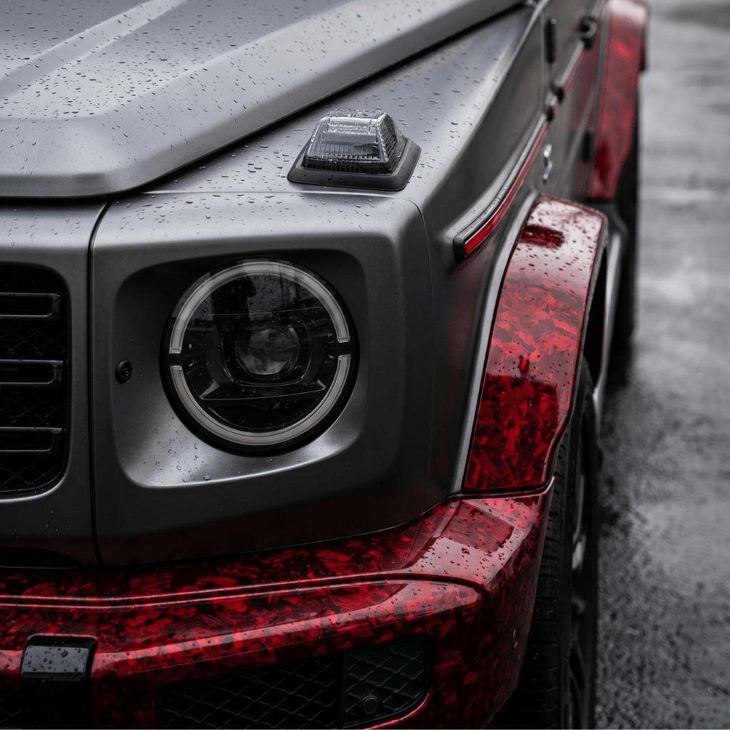 Close-up of the front left corner of a luxury SUV featuring a matte grey hood and Red Forged Carbon Graphite Self-Healing Nano-Coated PPF fender, highlighting the headlight, turn signal, and raindrops on the surface.