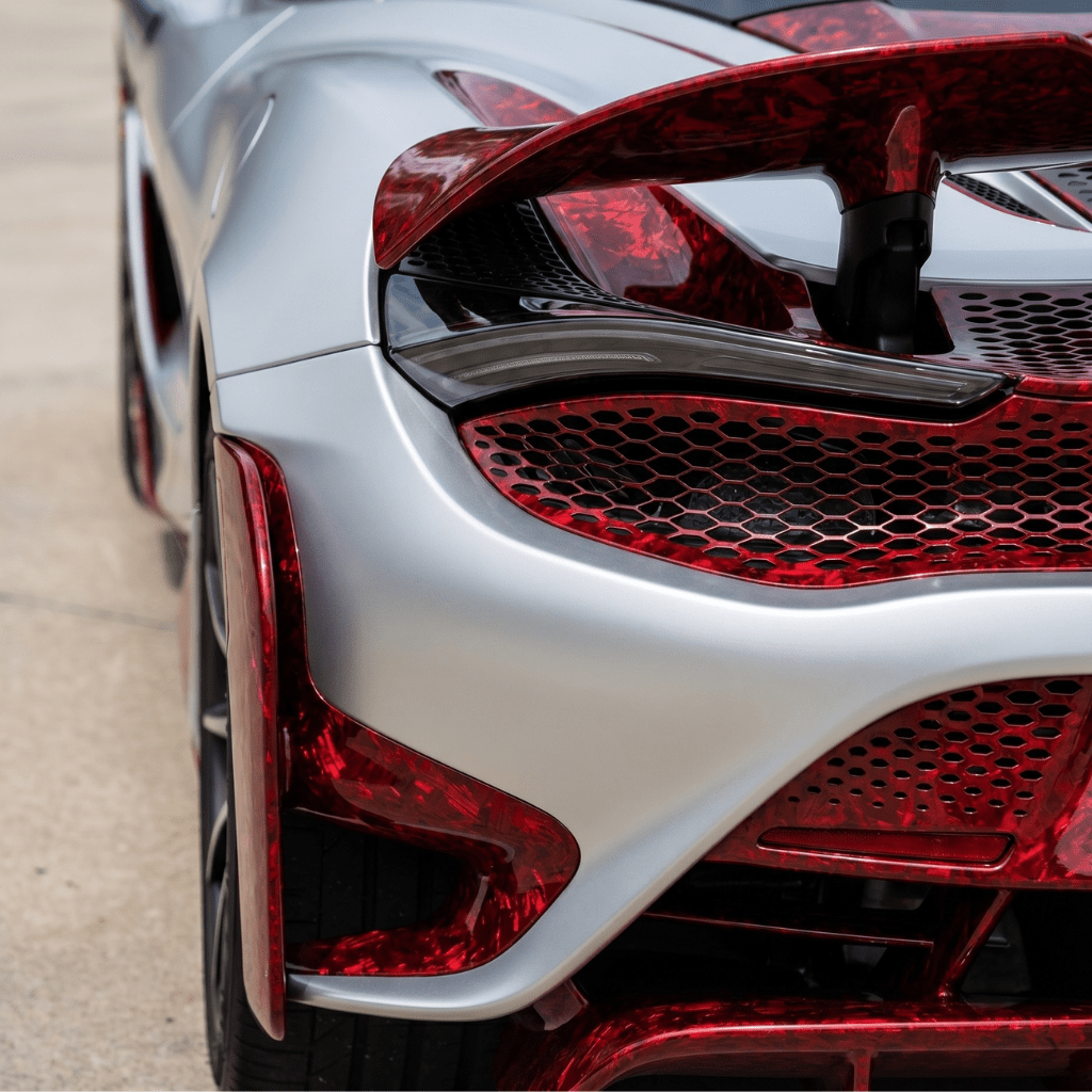 Close-up of the rear end of a silver sports car with sharp lines, unique rear lights and spoiler, featuring Red Forged Carbon Graphite Self-Healing Nano-Coated PPF accents. The background shows a blurred outdoor surface.