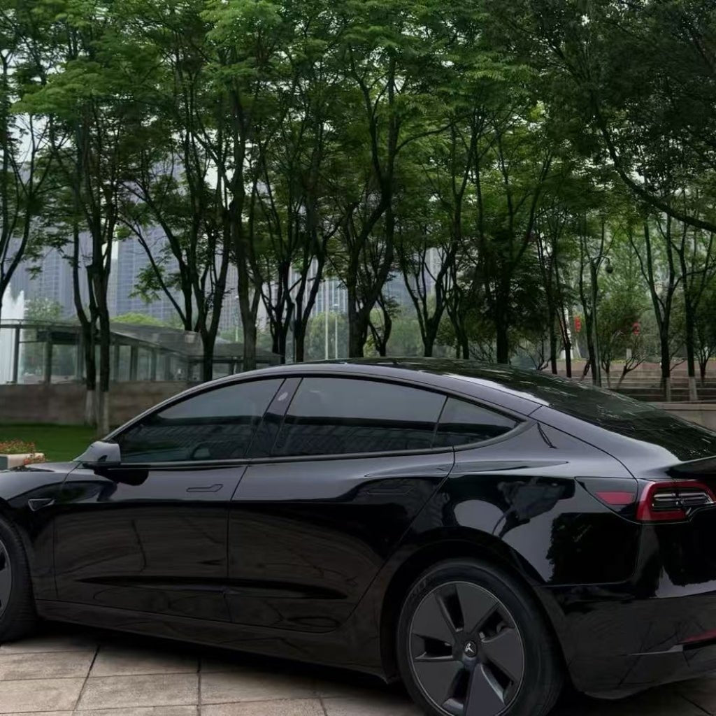 A black Tesla Model 3 with Shell Black Glossy SelfRepair Nano-Coated PPF is parked on a paved area, surrounded by tall green trees and a cityscape in the background.