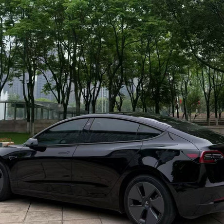 A black Tesla Model 3 with Shell Black Glossy SelfRepair Nano-Coated PPF is parked on a paved area, surrounded by tall green trees and a cityscape in the background.