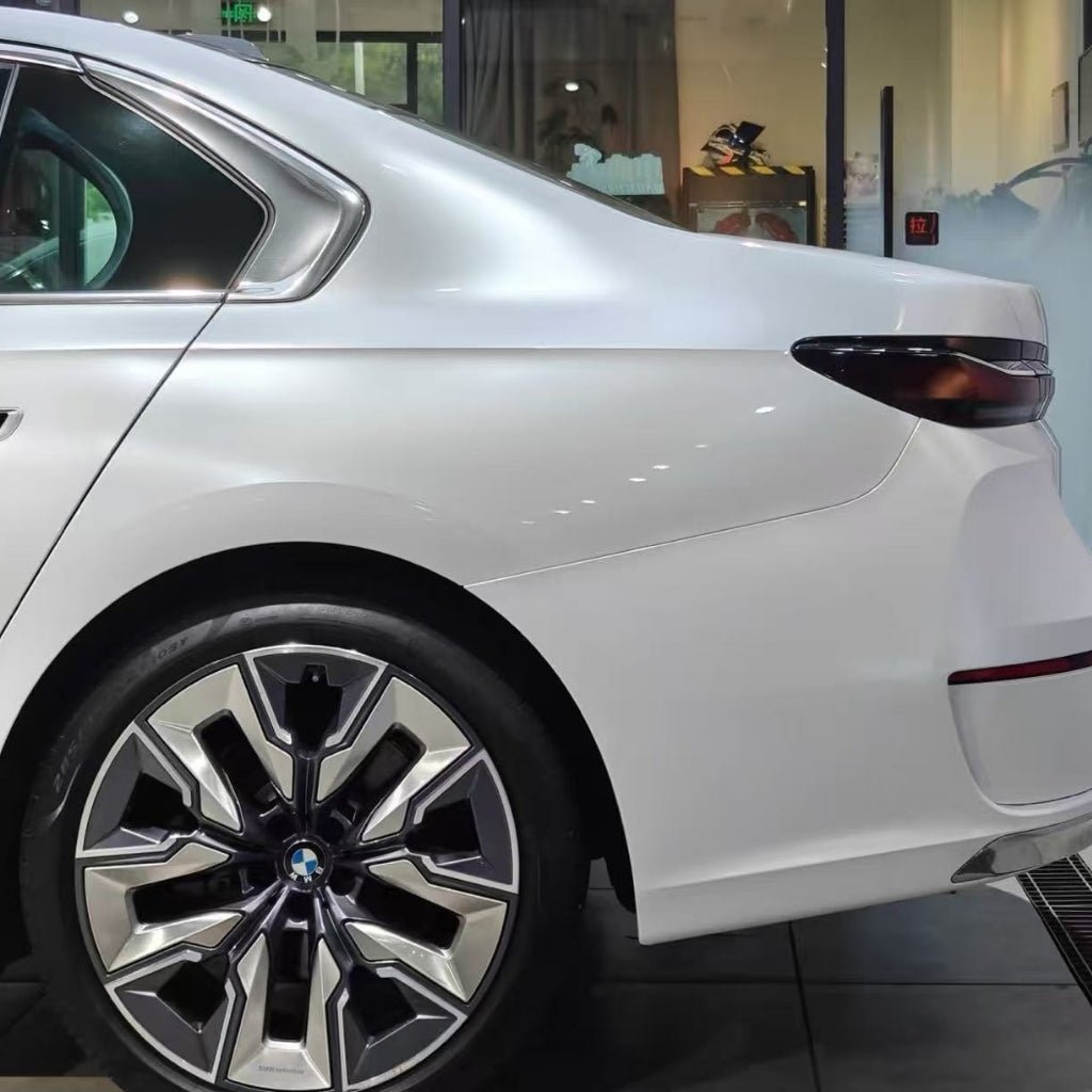 The image shows the rear of a white BMW sedan with Shell White Glossy SelfRepair Nano-Coated PPF, highlighting the back door, rear fender, and a large geometric alloy wheel. Glass walls and vivid reflections fill the background.