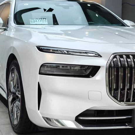 Close-up of a white luxury car with Shell White Glossy SelfRepair Nano-Coated PPF on the front left, highlighting the sleek headlight, chrome grille, and wheel. A paper sign is visible on the dashboard through the windshield.