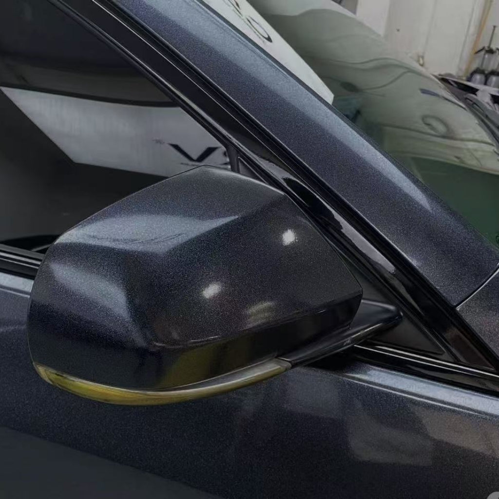 A close-up of a metallic gray car’s side mirror shielded by Sparkle Black Matte SelfRepair Nano-Coated Nexthor™ PPF and a partially open window, with reflections on its shiny surface and a softly blurred indoor background.