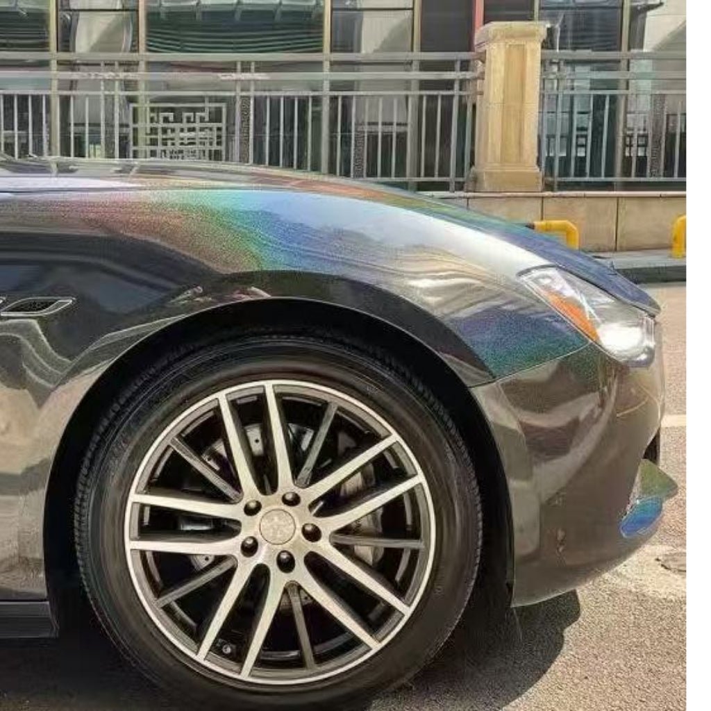 A close-up of the front left side of a dark-colored car with a rainbow sheen on the hood, featuring Stellar Black Glossy SelfRepair Nano-Coated PPF, parked on a street with a metal fence and building in the background.