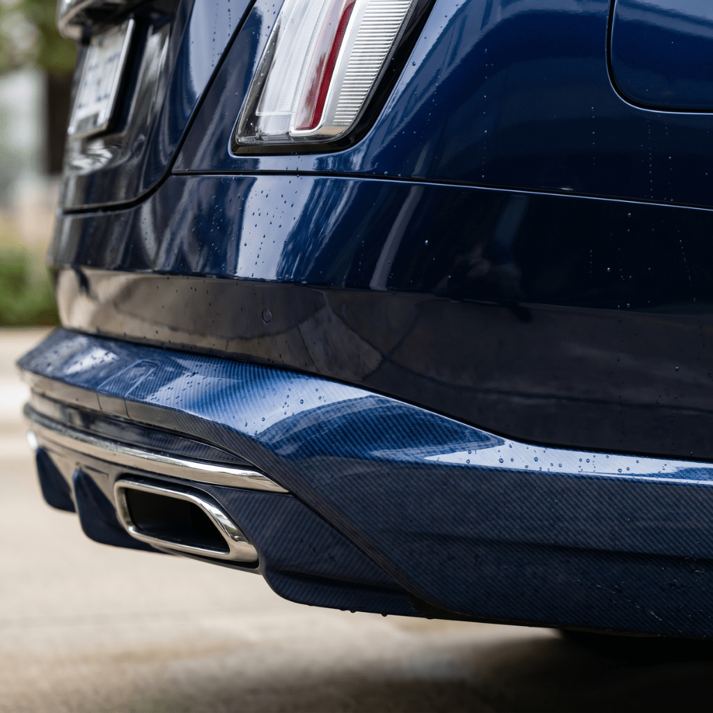 Close-up of a dark blue luxury car’s rear with chrome exhaust tips and carbon fiber details, protected by Transparent Carbon Fiber Self-Healing Nano-Coated PPF, its raindrop-covered surface shining near a light-colored building.
