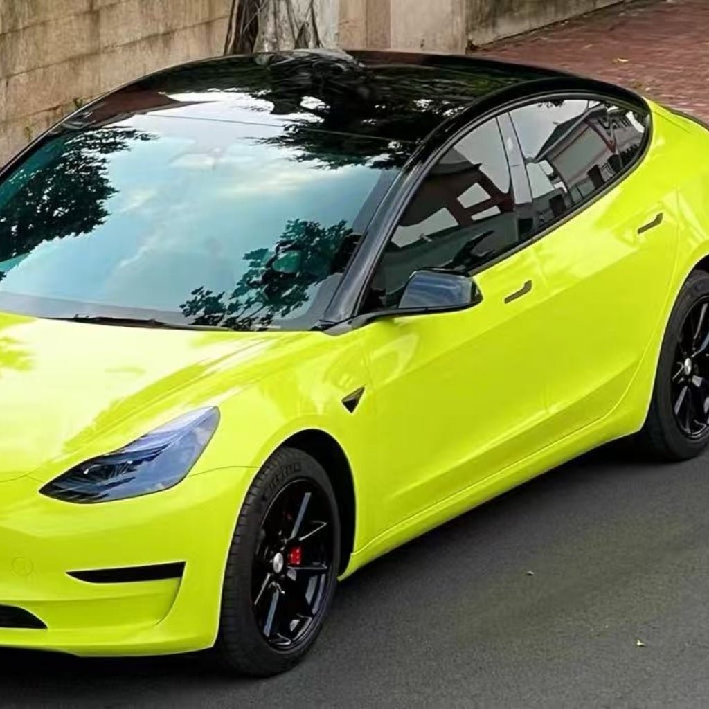 A Tesla sedan in bright yellow-green, with a black roof and wheels, is wrapped in São Paulo Glossy SelfRepair Nano-Coated PPF and parked by a stone wall. The car’s windshield and roof reflect nearby trees and clouds.