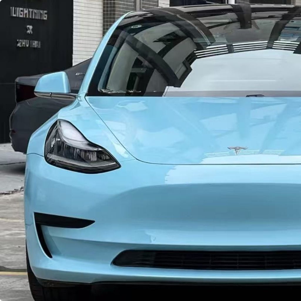 A Sea Breeze Blue Tesla, protected by Glossy SelfRepair Nano-Coated PPF, is parked outdoors near a building with the front and part of the side visible. Reflections of the surroundings appear on its hood and windshield.