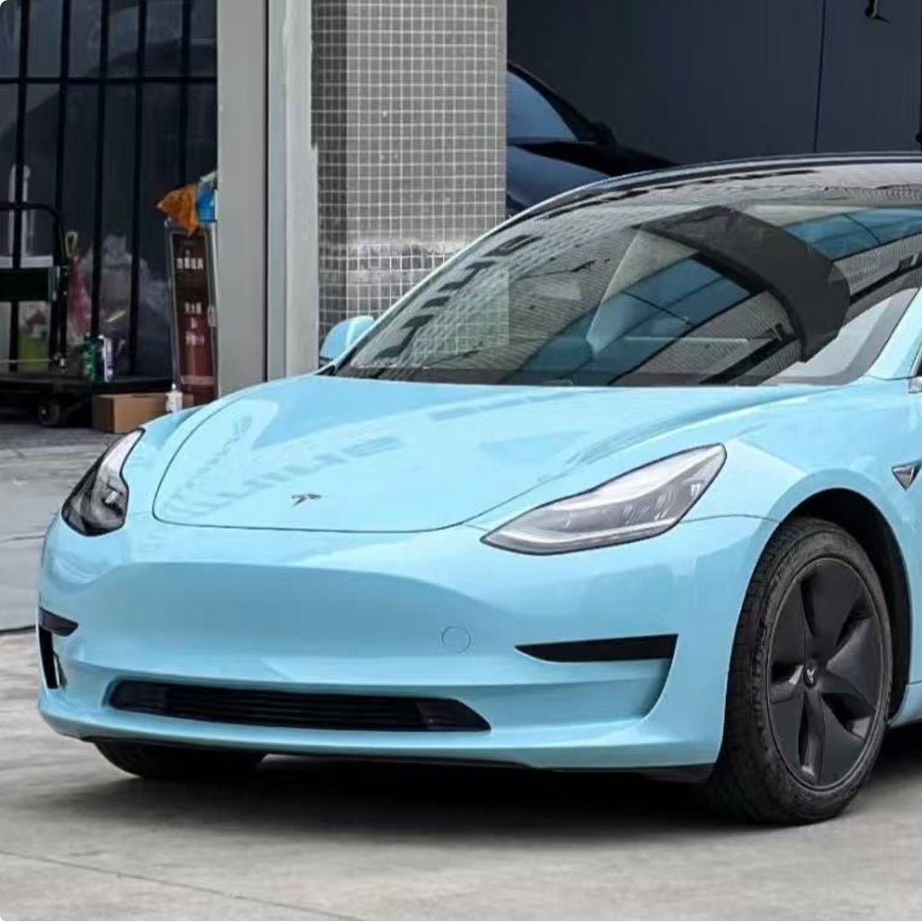 A light blue Tesla Model 3 with sleek headlights and black aero wheels is parked outdoors near a modern building, featuring Sea Breeze Blue Glossy SelfRepair Nano-Coated PPF for advanced paint protection.