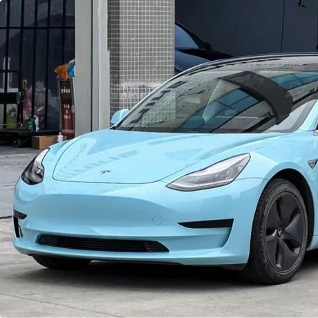 A light blue Tesla Model 3 with sleek headlights and black aero wheels is parked outdoors near a modern building, featuring Sea Breeze Blue Glossy SelfRepair Nano-Coated PPF for advanced paint protection.