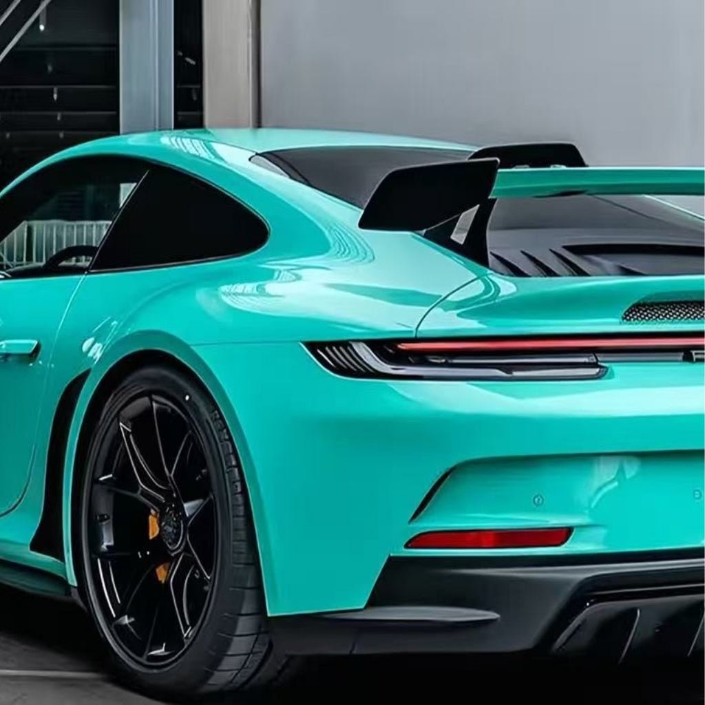A sports car in Tiffany Blue Glossy SelfRepair Nano-Coated PPF with a large rear spoiler and black wheels is parked by a modern building, its sleek lines protected by this premium paint protection film.