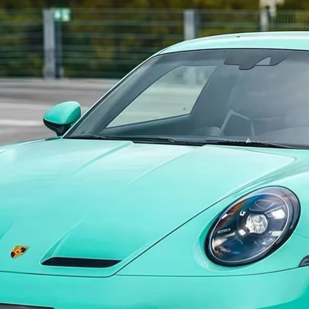A close-up shows the front of a turquoise Porsche featuring Tiffany Blue Glossy SelfRepair Nano-Coated PPF on its hood, left headlight, windshield, and side mirror. The car is parked outdoors with a blurred fence and greenery in the background.