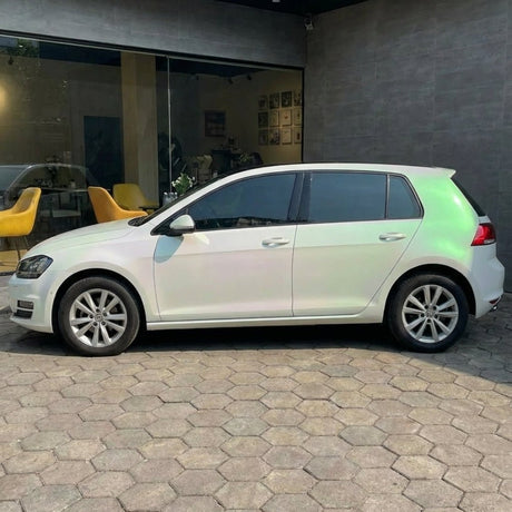 A white hatchback featuring White Diamond Pine Green Glossy SelfRepair Nano-Coated PPF is parked on a hexagonal stone driveway in front of a glass-walled building with yellow chairs inside.