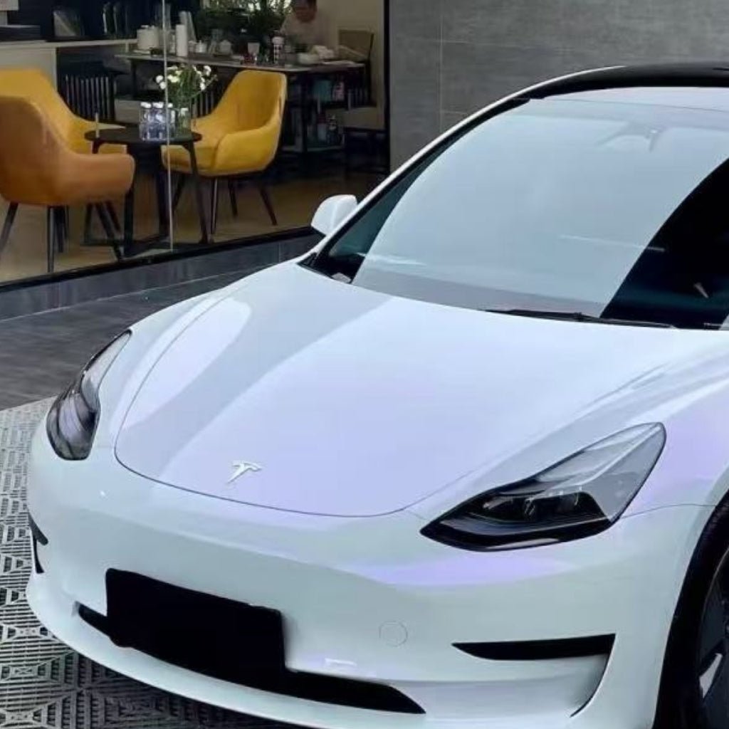 A white Tesla with White Diamond Purple Glossy SelfRepair Nano-Coated PPF is parked by a glass-walled room with yellow chairs, a dining table, and plants. The image highlights the car's sleek front and side with its glossy protective finish.