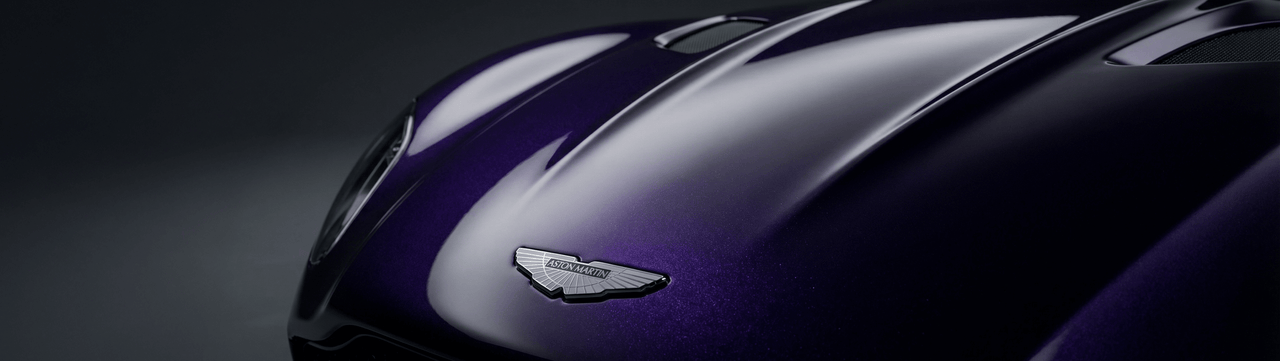 Close-up of a glossy, deep purple car hood featuring the Aston Martin emblem, with smooth curves and reflections visible under soft lighting.