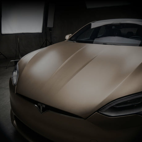 A close-up of Tesla wrapped in brown Nexthor PPF in a studio with soft lighting and photo equipment in the background. The focus is on the hood and front left headlight.