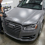 Inside a garage, a person uses a squeegee to apply Transparent Matte SelfRepair Nano-Coated PPF to the hood of a silver Audi S8, with cleaning supplies visible on a table in the background.