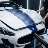 A person applies Transparent Matte SelfRepair Nano-Coated PPF to the hood of a white Maserati sports car with blue racing stripes in a detailing workshop.