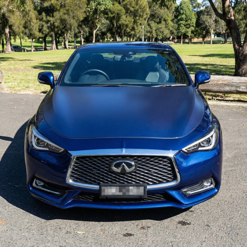 A blue Infiniti sedan with Transparent Matte Sparkle SelfRepair Nano-Coated PPF is parked on pavement by a park with green grass, trees, and sunlight in the background, viewed from the front.