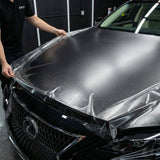 A person is applying Transparent Matte Sparkle SelfRepair Nano-Coated PPF to the hood of a black Lexus in a garage, carefully smoothing the protective film to ensure full coverage.