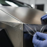 Wearing blue gloves, a person carefully applies or removes Transparent Matte Sparkle SelfRepair Nano-Coated PPF from a vehicle’s angular metallic surface, using self-healing nanotechnology for advanced paint protection.