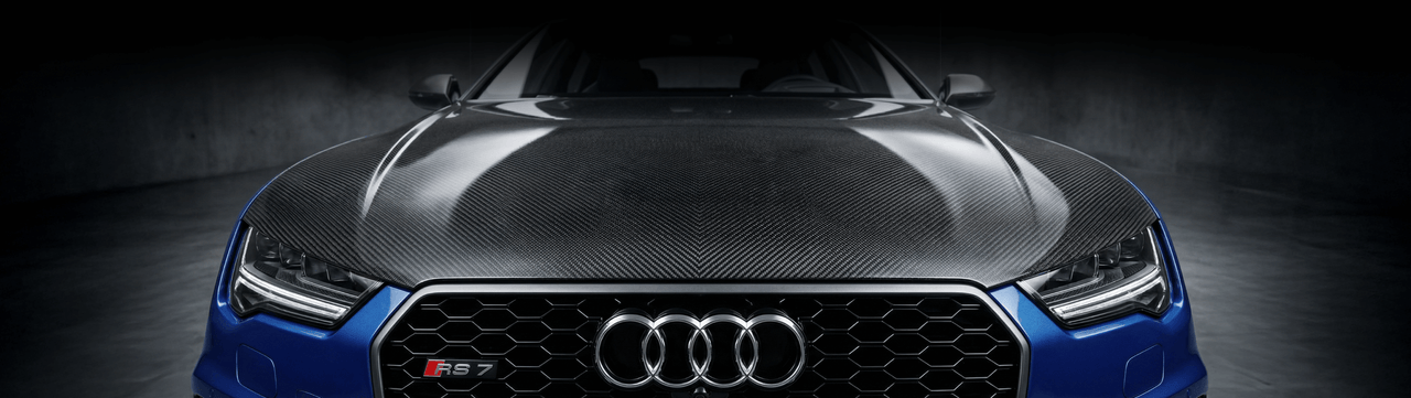 Close-up front view of a blue Audi RS7 with a carbon fiber hood, featuring the Audi logo and honeycomb grille, set against a dark, blurred background.