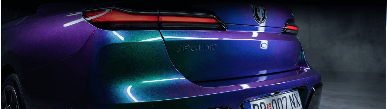 Rear view of a car with an iridescent purple-green paint job, displaying sleek LED tail lights, the word “NEXTHOR” embossed on the trunk, and a European license plate in an indoor setting.