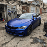 A BMW sports car in Satin Liquid Metallic Blue SelfRepair Nano-Coated PPF is parked in a rundown, graffiti-marked garage with maintenance tools and spare tires scattered amid worn industrial surroundings.