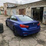 A matte blue BMW coupe with Satin Liquid Metallic Blue SelfRepair Nano-Coated PPF is parked in a cracked, weathered lot near old brick garages covered in graffiti, with stacked tires and an open door leading to a dimly lit workspace inside.