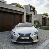 A silver Lexus wrapped in Satin Ceramic White SelfRepair Nano-Coated PPF is parked on a cobblestone driveway, framed by modern concrete homes with wooden garage doors and landscaped greenery.