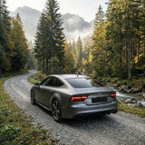 A Satin Liquid Metallic Gray Audi with SelfRepair Nano-Coated PPF is parked on a gravel road that winds through misty pine forests with a stream running alongside.