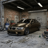 A matte bronze BMW sports car, wrapped in Satin Gold Green SelfRepair Nano-Coated PPF, sits in a gritty urban garage with graffiti, scattered tools and tires. Overhead lights enhance its luxurious finish and unique satin gold green look.