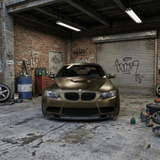 A BMW wrapped in Satin Gold Green SelfRepair Nano-Coated PPF is parked in an urban garage with exposed brick walls, tools, car parts, graffiti, and a metal shutter door in the background.