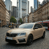 A Škoda sedan in Satin Twilight Gold, protected by Satin Twilight Gold SelfRepair Nano-Coated PPF, stands on a cobblestone street in a European city surrounded by historic and modern buildings with public transport passing by.