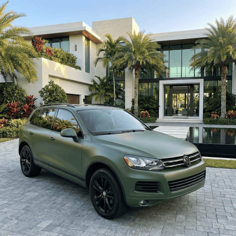 A Volkswagen SUV wrapped in Satin Army Green SelfRepair Nano-Coated PPF is parked on a stone driveway before a modern, luxurious house with palm trees and lush landscaping.