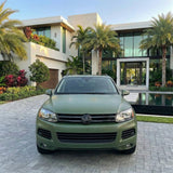 A Volkswagen SUV in Satin Army Green SelfRepair Nano-Coated PPF is parked on a stone driveway before a modern glass-fronted house, surrounded by palm trees and lush landscaping.