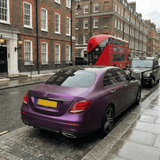 A Mercedes-Benz sedan wrapped in Satin Black Pinot Purple SelfRepair Nano-Coated PPF is parked on a wet city street near a black taxi and red double-decker bus. Brick buildings with white trim stand in the cloudy background.