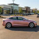 A pink Tesla sedan wrapped in Satin Rose Gold SelfRepair Nano-Coated PPF is parked in a shopping center lot on a sunny day, with storefronts and other cars in the background.