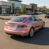 A Tesla sedan with Satin Rose Gold SelfRepair Nano-Coated PPF is parked in a sunny, empty shopping center lot, with retail stores and trees in the background.