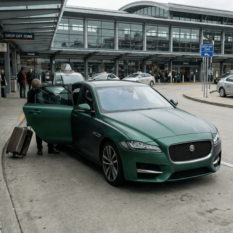 A traveler with a suitcase enters a green Jaguar sedan protected by Deep Emerald Satin Self-Healing Nano-Coated PPF, ensuring ochrana laku, at an airport drop-off zone as taxis and travelers pass the modern terminal.