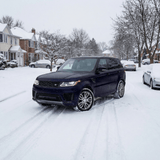 A dark SUV with Satin Midnight Steel SelfRepair Nano-Coated PPF glides down a snow-covered suburban street, bordered by houses, parked cars, and bare trees on a wintry day.