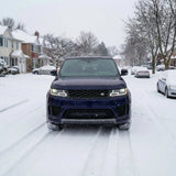 A black SUV with headlights on, protected by Satin Midnight Steel SelfRepair Nano-Coated PPF, is parked on a snowy residential street with houses and trees on both sides under a cloudy winter sky.