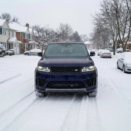 A black SUV with headlights on, protected by Satin Midnight Steel SelfRepair Nano-Coated PPF, is parked on a snowy residential street with houses and trees on both sides under a cloudy winter sky.