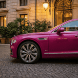 A bright pink luxury Bentley, protected by Gem Red Glossy SelfRepair Nano-Coated PPF, is parked on a cobblestone street before an elegant building with large windows, potted plants, and warm outdoor lighting.