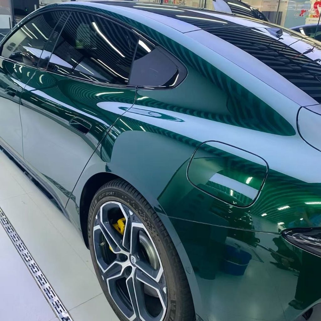 A shiny green electric car in Agate Green Liquid Metallic Glossy SelfRepair Nano-Coated PPF is seen from the side, displaying its rear door, sleek lines, dark tinted windows, and alloy wheels with yellow brake calipers under bright indoor lights.