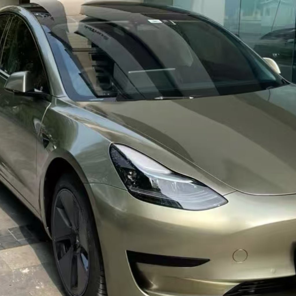 An Alpine Green Liquid Metallic Glossy SelfRepair Nano-Coated PPF-wrapped Tesla Model 3 is parked on a stone driveway beside a glass building, its shiny surface reflecting the nearby surroundings.