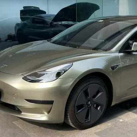 An Alpine Green Liquid Metallic Glossy SelfRepair Nano-Coated PPF Tesla Model 3 is parked outdoors, reflecting sunlight. Another car with its hood open appears in the background through glass windows.