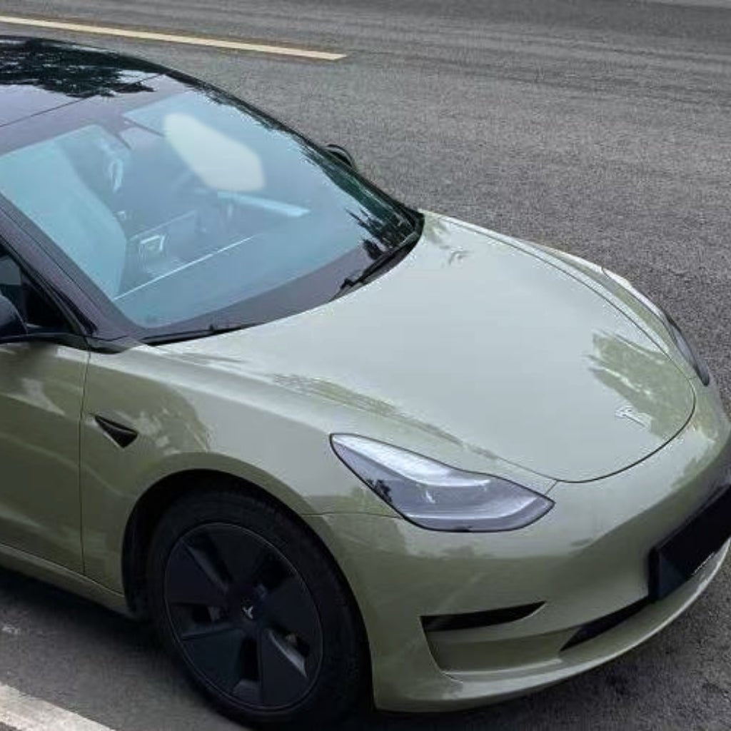 A Tesla sedan with black wheels is parked roadside, wrapped in Army Green Glossy SelfRepair Nano-Coated Nexthor™ PPF. The clear front and left side stand out, while greenery and the road complete the background scene.