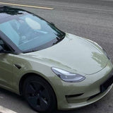 A Tesla sedan with black wheels is parked roadside, wrapped in Army Green Glossy SelfRepair Nano-Coated Nexthor™ PPF. The clear front and left side stand out, while greenery and the road complete the background scene.