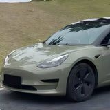 A Tesla Model 3 in light army green with black wheels, protected by Army Green Glossy SelfRepair Nano-Coated Nexthor™ PPF, is parked on a road before a grassy hillside.