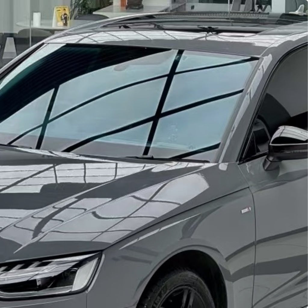 A close-up of a sleek car with Audi Nardo Gray Glossy SelfRepair Nano-Coated PPF, parked indoors. The windshield, hood, and side mirror shine with reflections from large windows on the protective film.